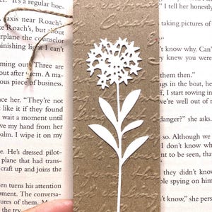 May include: A brown paper bookmark with a white floral design. The bookmark has a textured surface with embossed script and a twine string. The floral design is a stylized flower with a cluster of petals and a long stem.