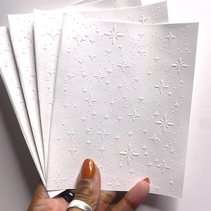 May include: A stack of white, textured paper cards with an embossed star pattern. The cards are held in a hand with a ring and orange nail polish. The star design includes various sizes of stars and dots.