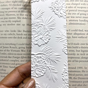 May include: White bookmark with an embossed pine cone and branch design. The bookmark has a hole at the top with a natural twine string. The bookmark is rectangular and is held over an open book.