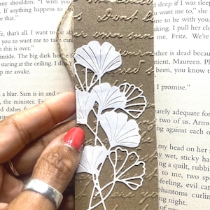 May include: A handmade bookmark with a brown background and white floral cutouts. The bookmark has a textured surface with embossed text. A natural twine is attached at the top for easy placement in a book. The bookmark is held by a person's hand.