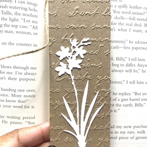May include: A rectangular brown paper bookmark featuring an embossed cursive script background and a die-cut white paper flower silhouette attached to the front. A thin jute twine loop is threaded through a hole at the top of the bookmark.