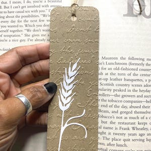 May include: A brown paper bookmark with a white wheat stalk design. The bookmark has a textured surface with embossed text and a hole at the top with a twine string. A unique accessory for book lovers.