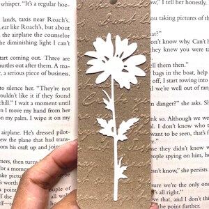 May include: A brown paper bookmark with a white daisy cut-out design. The bookmark has a textured surface and a twine string for hanging. The background shows printed text.