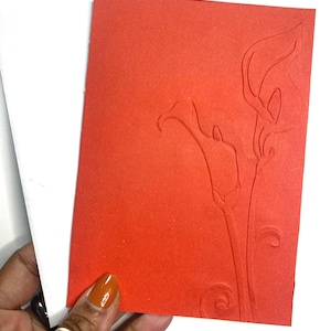 May include: A red greeting card with an embossed calla lily design. The card is held next to a white envelope. The calla lily design is a detailed floral pattern, adding a touch of elegance to the card.