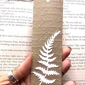 May include: A brown paper bookmark with a white fern cutout and embossed script. The bookmark is rectangular with a hole at the top for a twine string. The background is a book page.
