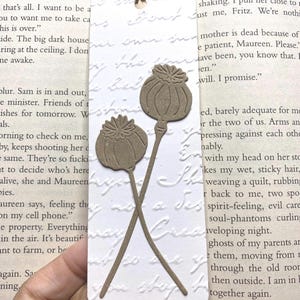 May include: A handmade bookmark featuring two brown poppy seed head cutouts. The bookmark is rectangular with embossed script and a twine hanger. The poppy seed heads are detailed and crossed, adding a touch of nature to the design.