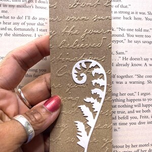 May include: A brown paper bookmark with embossed script and a white fern-like cutout. The bookmark has a string for easy placement in a book. The text on the bookmark is partially visible.