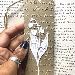 May include: A rectangular brown paper bookmark featuring an embossed cursive script pattern and a white die-cut lily of the valley flower silhouette attached to the center. A thin jute twine loop is threaded through a hole at the top edge.