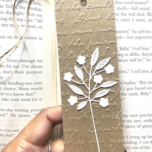 May include: A rectangular brown paper bookmark featuring an embossed cursive script texture across the surface. A white die-cut floral sprig with five small flowers and several leaves is attached to the center. A thin jute twine loop is tied at the top.