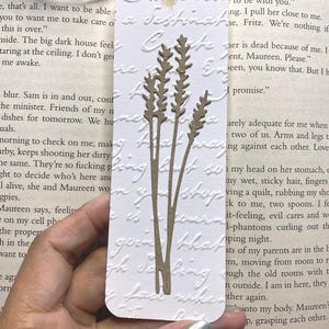 May include: A white bookmark with a brown floral design and a jute string. The bookmark has embossed script and a die-cut wheat stalk design. The bookmark is held over an open book.