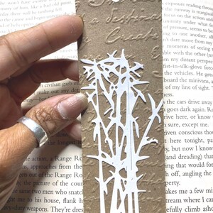 May include: A handmade bookmark with a white bamboo design cut-out against a textured brown background. The bookmark has a jute string for hanging. The text on the bookmark reads "A Destination Create".