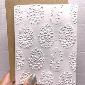 May include: A white embossed Easter card featuring floral and botanical designs within egg shapes. The card is held with a brown envelope. The card's texture and design are highlighted in this close-up shot.