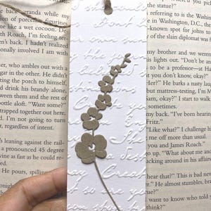 May include: A white bookmark with embossed script and a brown floral design. The bookmark is rectangular with a rounded bottom and a twine loop at the top. The floral design is a stem with several flower shapes.