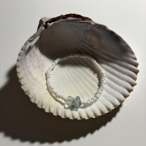 May include: A white beaded bracelet with three light blue gemstone accents, displayed in a seashell.
