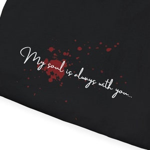 Peut inclure: T-shirt noir plié, avec l'inscription blanche "My soul is always with you..." et des éclaboussures rouges. Le texte est en anglais. Fond blanc.