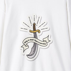 Pode incluir: Camisola branca com um gráfico de uma espada com o texto "Body in abyss, heart in paradise" impresso nela.