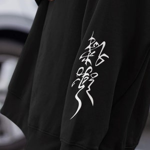 Pode incluir: Camisola de moletom preta oversized com um design gráfico branco na manga. O design apresenta uma imagem estilizada de uma árvore com a palavra "Paz" escrita em caracteres japoneses.