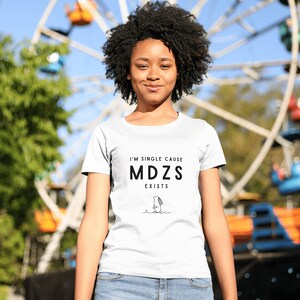 K&ouml;nnte beinhalten: Eine Person tr&auml;gt ein wei&szlig;es T-Shirt mit dem Text "I'm single cause MDZS exists" und einer Schwarzwei&szlig;illustration einer Person. Die Person steht vor einem Riesenrad.