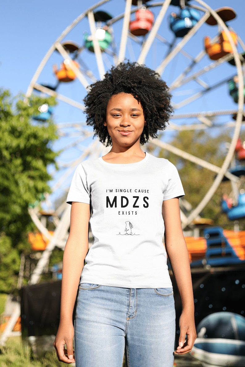 K&ouml;nnte beinhalten: Eine Person tr&auml;gt ein wei&szlig;es T-Shirt mit dem Text "I'm single cause MDZS exists" und einer Schwarzwei&szlig;illustration einer Person. Die Person steht vor einem Riesenrad.