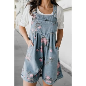 May include: Pale blue denim dungaree shorts featuring a pink and green floral print. The shorts have a bib front with pockets and adjustable straps. Worn over a white t-shirt.