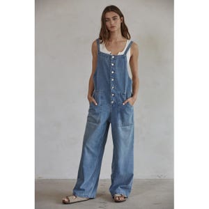 Op de afbeelding: Lichtblauwe denim overall met een knoopsluiting aan de voorkant en wijde pijpen. De overall heeft twee voorzakken en verstelbare bandjes. Er wordt een witte tanktop onder gedragen. De persoon draagt sandalen.
