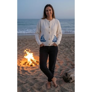 May include: Cream-colored cardigan with two blue sailboat appliques on the front. The button-down sweater is paired with black jeans. A bonfire burns on the beach in the background.