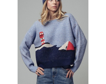 May include: A light blue long-sleeve knit sweater featuring a dark blue bottom section with a graphic design of a red and white lighthouse and a red and white sailboat. The sweater is paired with a dark blue knit beanie and blue denim jeans.