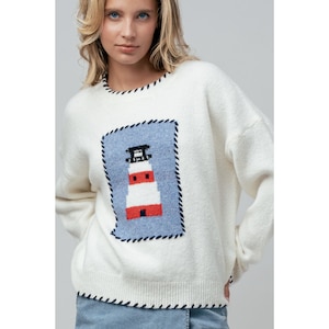 Könnte beinhalten: Cremefarbener Pullover mit Leuchtturm-Design. Der Pullover hat ein blaues Quadrat mit einer Leuchtturmillustration in Rot, Weiß und Schwarz. Der Ausschnitt und die Manschetten haben eine schwarz genähte Bordüre.