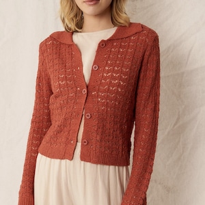 May include: A rust-colored, open-weave cardigan with a collared neckline and button-down front. The long-sleeved sweater has a textured pattern and is paired with cream-colored pants. The cardigan is a warm, earthy tone, suitable for layering.