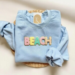 May include: A light blue sweatshirt with the word "BEACH" in colorful, textured letters. The letters are pink, yellow, and green. The sweatshirt is folded on a woven mat.