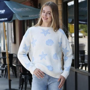 May include: A white sweater with a crew neck, featuring a pattern of light blue starfish and seashells. The sweater has long sleeves and is made of a soft, textured material. The person is wearing blue jeans.