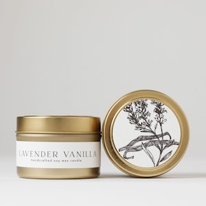 May include: A gold tin candle with a white label that reads "Lavender Vanilla handcrafted soy wax candle". The tin has a black and white illustration of lavender and vanilla beans.