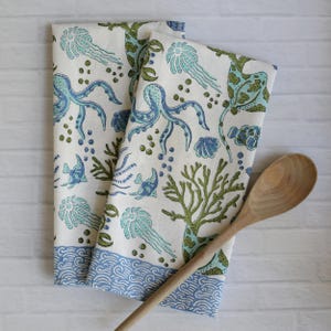 May include: Two white kitchen towels with a blue and green sea life pattern, including octopuses, jellyfish, and coral. The towels have a blue wave border. A wooden spoon rests on the side.