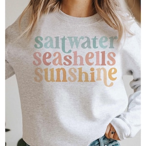 以下が含まれることがあります： パステルカラーで「saltwater seashells sunshine」の文字がプリントされたライトグレーのスウェットシャツ。テキストは縦に積み重ねられ、各単語が別の行にあります。スウェットシャツはカジュアルで快適なスタイルです。