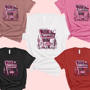 May include: Five different color t-shirts with a retro-style graphic of a neon sign that reads "Welcome to Road On Smutville". The sign is pink and white with a black outline. The t-shirts are pink, white, black, red, and heather grey.