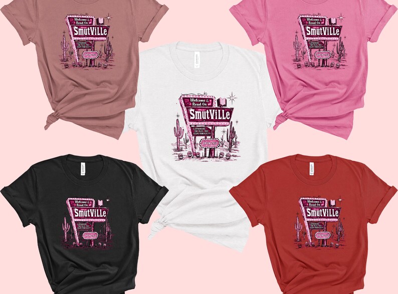 May include: Five different color t-shirts with a retro-style graphic of a neon sign that reads "Welcome to Road On Smutville". The sign is pink and white with a black outline. The t-shirts are pink, white, black, red, and heather grey.