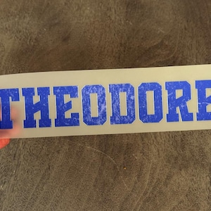 May include: A rectangular, semi-transparent transfer sheet featuring the name THEODORE in bold, block-style capital letters. The lettering has a vibrant blue color with a distressed, weathered texture throughout each character.