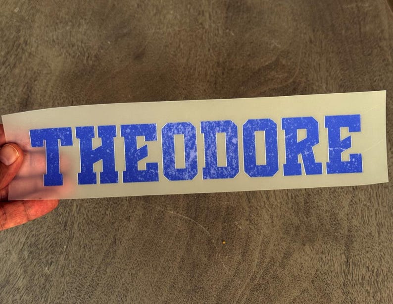 May include: A rectangular, semi-transparent transfer sheet featuring the name THEODORE in bold, block-style capital letters. The lettering has a vibrant blue color with a distressed, weathered texture throughout each character.