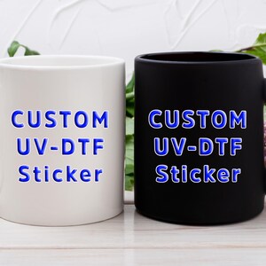 Puede incluir: Una taza de cer&aacute;mica blanca y una taza de cer&aacute;mica negra mate, cada una con el texto azul "CUSTOM UV-DTF Sticker" impreso en el frente. Ambas tazas tienen un asa est&aacute;ndar y un cuerpo cil&iacute;ndrico, mostrando la aplicaci&oacute;n de la calcoman&iacute;a.