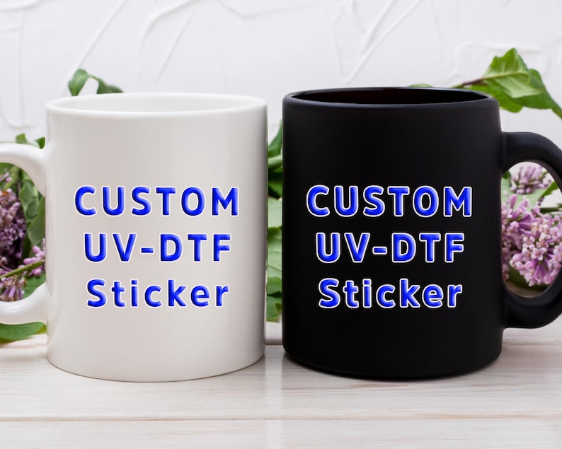 Puede incluir: Una taza de cer&aacute;mica blanca y una taza de cer&aacute;mica negra mate, cada una con el texto azul "CUSTOM UV-DTF Sticker" impreso en el frente. Ambas tazas tienen un asa est&aacute;ndar y un cuerpo cil&iacute;ndrico, mostrando la aplicaci&oacute;n de la calcoman&iacute;a.