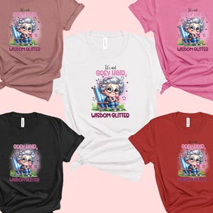 Op de afbeelding: Vijf verschillende gekleurde T-shirts met een afbeelding van een cartoon oma die op een stoel zit met de tekst "Het is geen grijs haar, wijsheid glinstert".