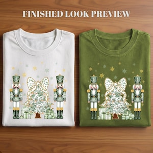 May include: A white t-shirt and an olive green t-shirt, each featuring a graphic of two nutcrackers flanking a Christmas tree with a large bow and wrapped gifts, accented by gold and green snowflakes and the text FINISHED LOOK PREVIEW.