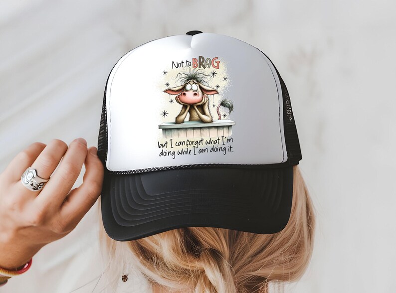 May include: A black and white trucker hat with a white front panel featuring a cartoon donkey graphic. The donkey is resting its chin on a ledge, with the text "Not to BRAG but I can forget what I'm doing while I am doing it."