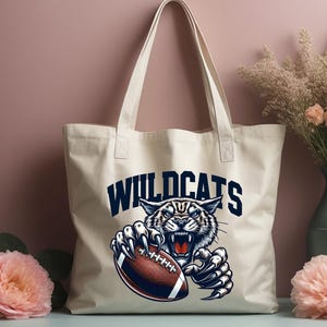 Może przedstawiać: Biała torba typu tote z pł&oacute;tna z niebiesko-białym nadrukiem przedstawiającym dzikiego kota trzymającego piłkę do futbolu. Tekst "WILDCATS" jest nadrukowany na niebiesko nad grafiką.