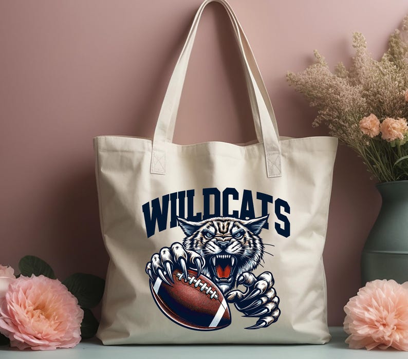 Może przedstawiać: Biała torba typu tote z pł&oacute;tna z niebiesko-białym nadrukiem przedstawiającym dzikiego kota trzymającego piłkę do futbolu. Tekst "WILDCATS" jest nadrukowany na niebiesko nad grafiką.