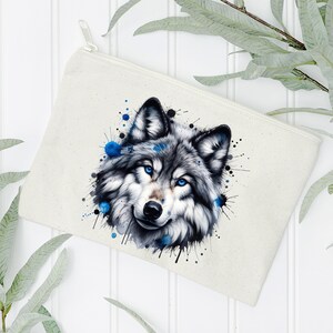 May include: A white canvas makeup bag with a watercolor-style illustration of a wolf's face. The wolf has blue eyes and is surrounded by blue and black paint splatters.