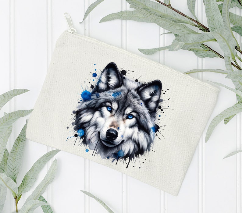 May include: A white canvas makeup bag with a watercolor-style illustration of a wolf's face. The wolf has blue eyes and is surrounded by blue and black paint splatters.