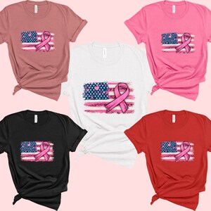 May include: Five t-shirts in different colors, each featuring a distressed American flag with a pink ribbon in the center. The ribbon is a symbol of breast cancer awareness.