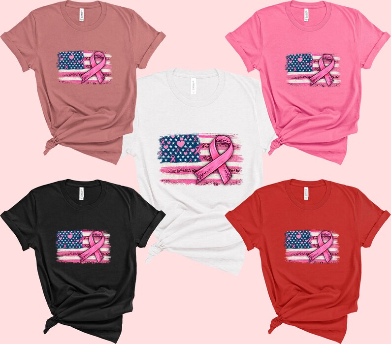 May include: Five t-shirts in different colors, each featuring a distressed American flag with a pink ribbon in the center. The ribbon is a symbol of breast cancer awareness.