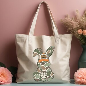 May include: A white canvas tote bag featuring a camouflage bunny rabbit wearing a hat that says "Get Lucky". The bag has two handles and is resting on a pink surface.